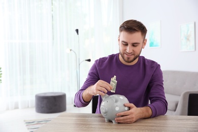 Young man putting money into piggy bank at table indoors. Space for text
