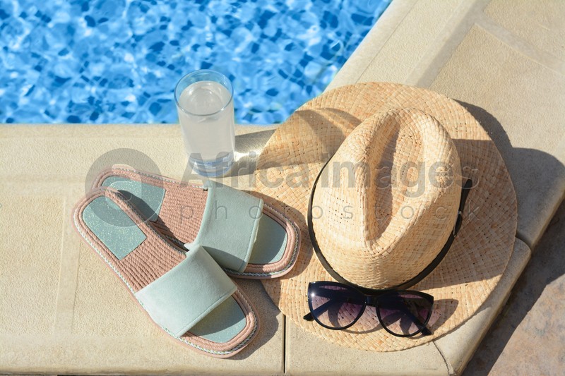 Stylish sunglasses, slippers, straw hat and glass of water at poolside on sunny day. Beach accessories