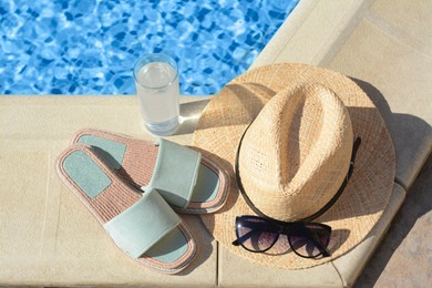 Photo of Stylish sunglasses, slippers, straw hat and glass of water at poolside on sunny day. Beach accessories
