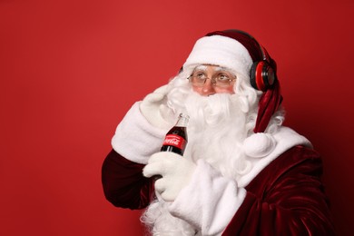 MYKOLAIV, UKRAINE - JANUARY 18, 2021: Santa Claus holding Coca-Cola bottle and listening to music with headphones on red background