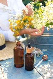 Woman with flowers near table outdoors, focus on bottles of chamomile essential oil
