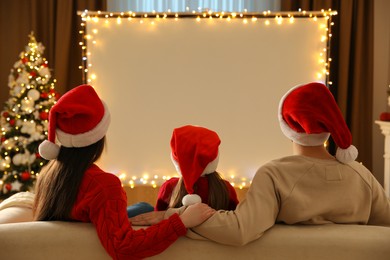 Family watching movie on projection screen in room decorated for Christmas, back view. Home TV equipment