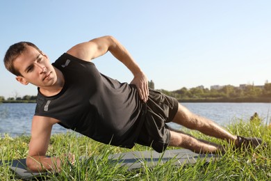 Sporty man doing side plank exercise on green grass near river