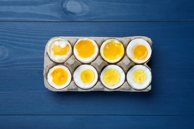 Photo of Boiled chicken eggs of different readiness stages in carton on blue wooden table, top view