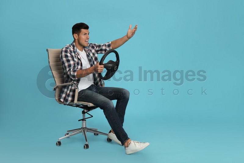 Emotional man on chair with steering wheel against light blue background. Space for text