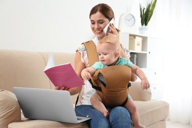 Woman with her son in baby carrier and book talking on phone at home