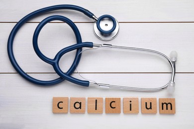 Photo of Word Calcium made of cubes with letters and stethoscope on white wooden table, top view
