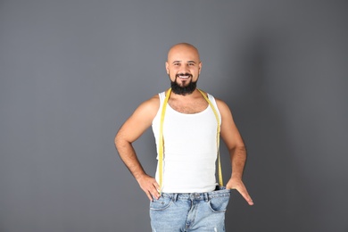Overweight man with measuring tape wearing oversized pants on gray background