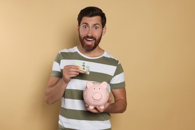 Excited man putting money into piggy bank on beige background