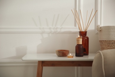 Photo of Oil reed diffuser and candles on wooden table near white wall, space for text. Interior decor