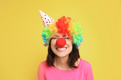 Joyful woman with rainbow wig, party hat and clown nose on yellow background. April fool's day