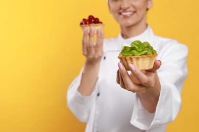 Photo of Happy professional confectioner in uniform with delicious tartlets on yellow background, closeup. Space for text