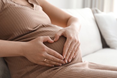 Pregnant young woman making heart with her hands on belly at home, closeup