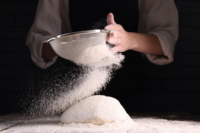 Photo of Woman sprinkling flour over dough at wooden table on dark background, closeup