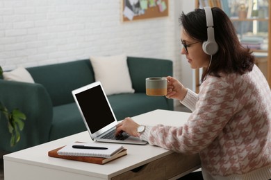 Woman with modern laptop and headphones drinking tea while learning at table indoors