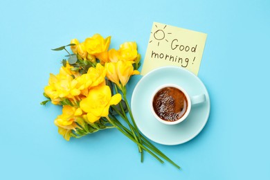 Cup of aromatic coffee, beautiful yellow freesias and Good Morning note on light blue background, flat lay