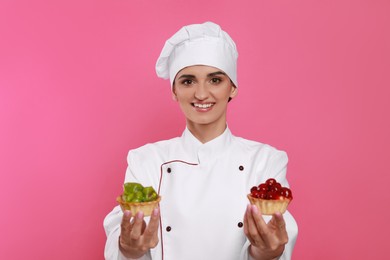 Happy professional confectioner in uniform with delicious tartlets on pink background