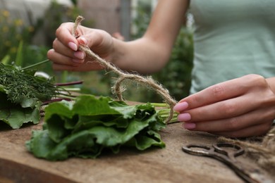 Woman tying bunch of fresh green leaves with twine at wooden table outdoors, closeup. Drying herbs