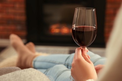 Woman with glass of wine relaxing near fireplace at home, closeup. Space for text