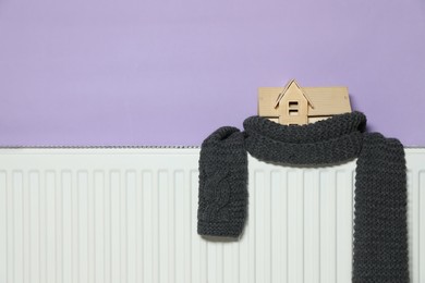 Wooden house model and knitted scarf on radiator near violet wall, space for text. Heating efficiency