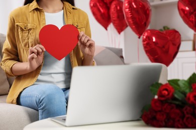Photo of Valentine's day celebration in long distance relationship. Woman holding red paper heart while having video chat with her boyfriend via laptop, closeup