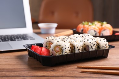 Tasty sushi rolls with shrimps in box near laptop on wooden table indoors