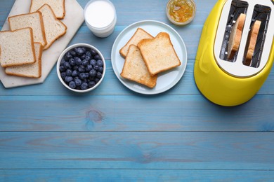 Yellow toaster with roasted bread, glass of milk, blueberries and jam on light blue wooden table, flat lay. Space for text
