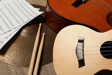 Ukulele, acoustic guitar, drumsticks, cymbal and note sheets on wooden background, closeup. Musical instruments