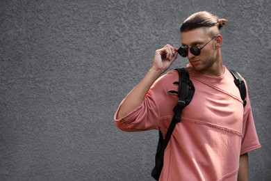 Handsome young man in stylish sunglasses and backpack near grey wall, space for text