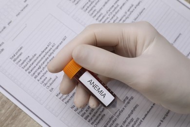 Doctor holding test tube with blood sample and label Anemia over medical form, above view