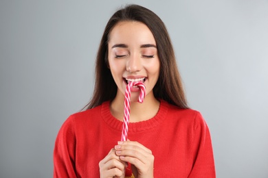 Young woman in Christmas sweater eating candy cane on grey background