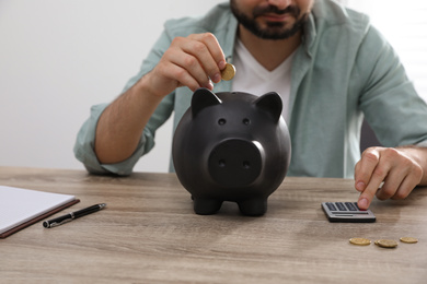Man with calculator putting coin into piggy bank at wooden table, closeup