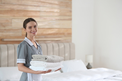 Young chambermaid holding stack of fresh towels in bedroom. Space for text