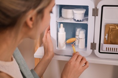 Woman with face roller taking cosmetic product from mini fridge indoors, closeup