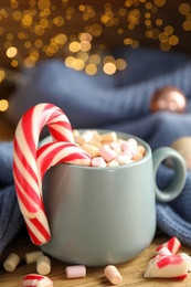 Cup of tasty cocoa with marshmallows and Christmas candy canes on wooden table against blurred festive lights