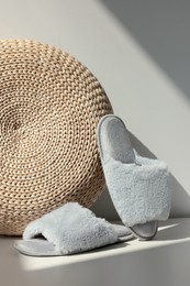 Pair of soft slippers and wicker pouf on floor near white wall