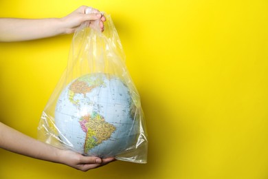 Woman holding globe in plastic bag against yellow background, closeup. Space for text. Environmental conservation