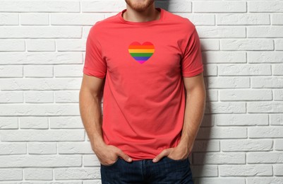 Young man wearing t-shirt with image of heart shaped LGBT pride flag near white brick wall
