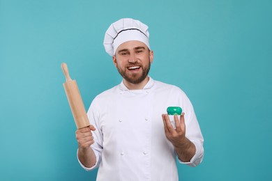 Photo of Happy professional confectioner in uniform with delicious cake and rolling pin on light blue background
