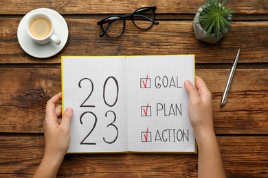Woman holding notebook with text 2023 Goal, Plan, Action at wooden table, top view. New Year resolutions