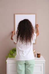 Woman hanging frame on pale rose wall over chest of drawers in room, back view