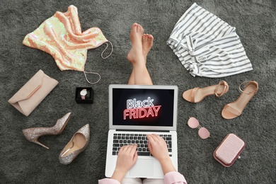 Woman surrounded by clothes and accessories using laptop with Black Friday announcement while sitting on grey carpet, top view