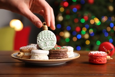 Woman taking Christmas macaron from plate at wooden table against blurred festive lights, closeup