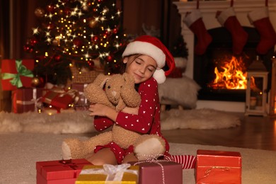 Cute child in Santa hat hugging toy bunny near Christmas gifts on floor at home