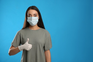 Woman in protective face mask and medical gloves showing thumb up gesture on blue background. Space for text