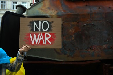 Woman wrapped in Ukrainian flag holding poster with words No War near broken military equipment on street, closeup