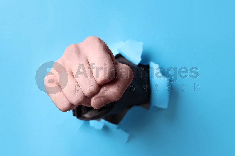 Photo of Businessman breaking through light blue paper with fist, closeup
