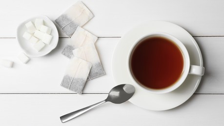Tea bags and sugar near cup of hot drink on white wooden table, flat lay