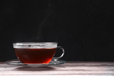 Photo of Glass cup of tea and saucer on wooden table against black background, space for text