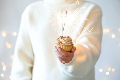 Woman holding cupcake with burning sparklers against blurred festive lights, closeup